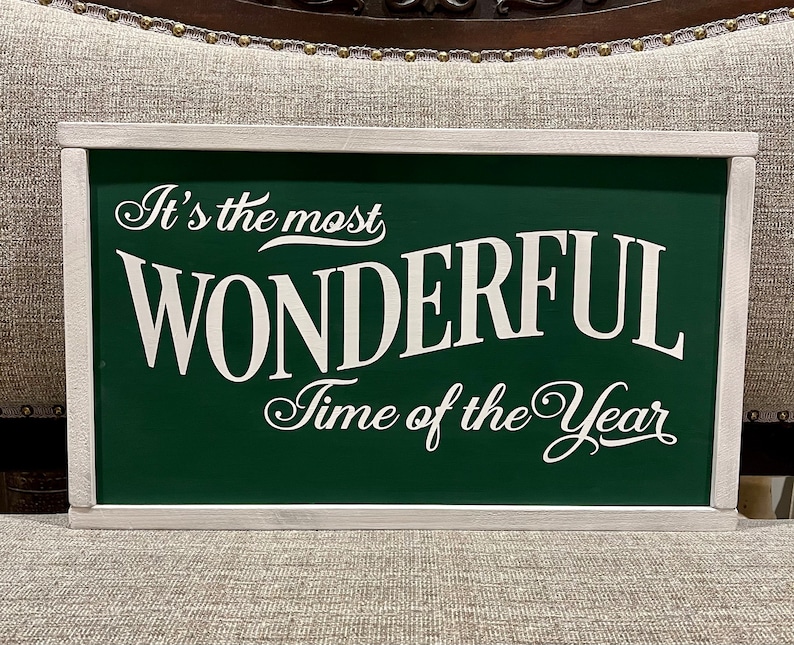 Puede incluir: Un letrero verde enmarcado en blanco con el texto "It's the most WONDERFUL Time of the Year" en fuente blanca cursiva.