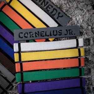 Może przedstawiać: Czarny stojak na pasy z nazwami "Kennedy" i "Cornelius Jr." na nim. Stojak zawiera kolekcję pas&oacute;w do sztuk walki w r&oacute;żnych kolorach, w tym biały, ż&oacute;łty, pomarańczowy, zielony, fioletowy, niebieski, brązowy i czarny.