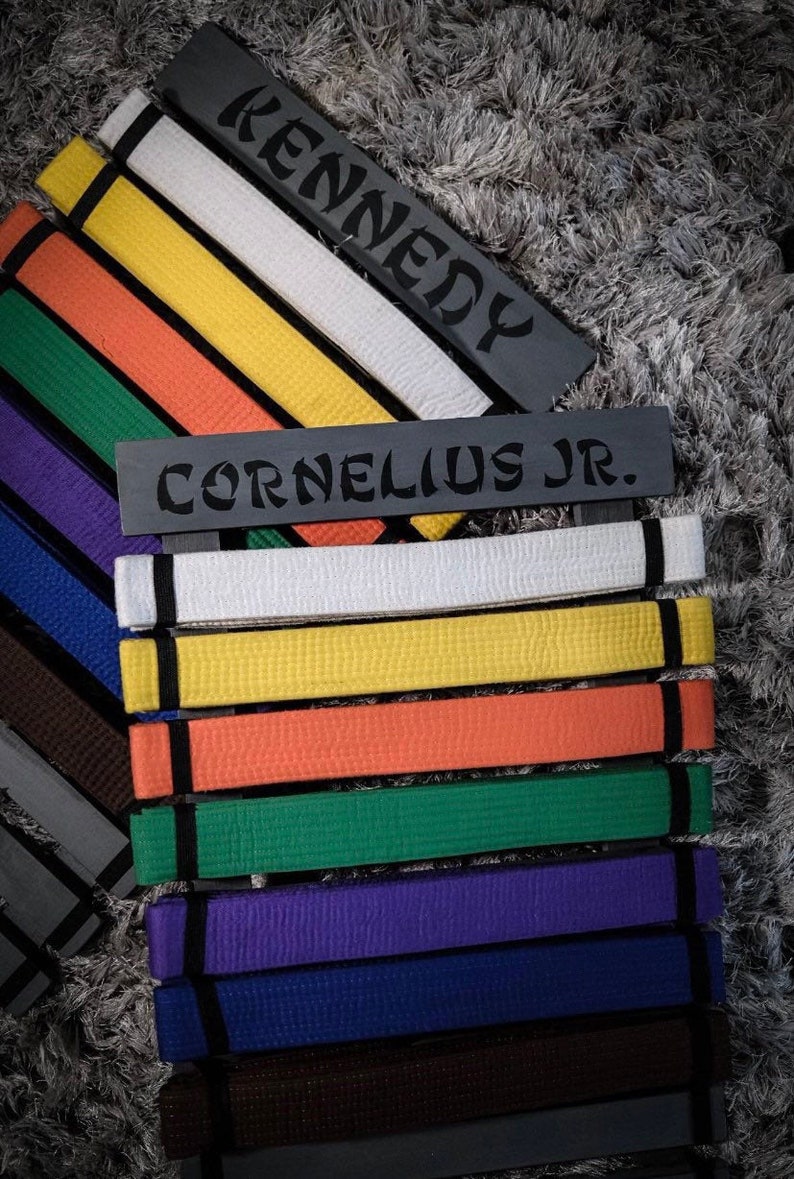 Może przedstawiać: Czarny stojak na pasy z nazwami "Kennedy" i "Cornelius Jr." na nim. Stojak zawiera kolekcję pas&oacute;w do sztuk walki w r&oacute;żnych kolorach, w tym biały, ż&oacute;łty, pomarańczowy, zielony, fioletowy, niebieski, brązowy i czarny.