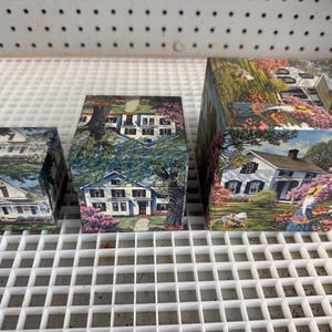 May include: Three square wooden boxes of varying sizes, each decorated with a printed scene of a white house surrounded by blooming pink flowers and green trees. The boxes feature a consistent landscape illustration across their exterior surfaces.
