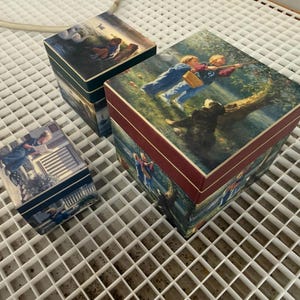 May include: Three square decorative boxes of varying sizes, each featuring a different printed illustration of children in outdoor settings. The largest box has a dark red border, while the two smaller boxes feature dark blue accents and edges.