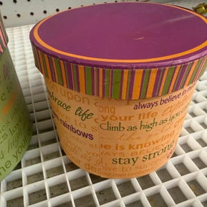 May include: A round cardboard storage box with a purple lid featuring an orange border. The sides are orange with vertical striped trim and printed text including phrases like embrace life, climb as high as you can, and stay strong in various fonts.