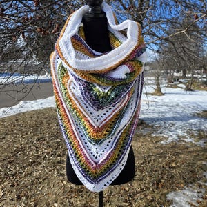 May include: A handmade crochet shawl in a triangular shape, displayed on a black mannequin. The shawl features a white border and a vibrant array of colors including yellow, green, purple, and pink. The background shows a winter scene with bare trees.