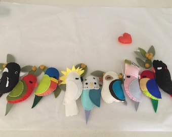 Könnte beinhalten: Eine Filzgirlande mit neun bunten Vögeln, darunter ein schwarz-weißer Vogel, ein roter Vogel, ein grün-gelber Vogel, ein weißer Vogel mit gelber Haube, ein blauer Vogel, ein weißer Vogel mit blauem Flügel, ein rosa Vogel, ein blau-gelber Vogel und ein schwarzer Vogel. Die Girlande ist mit grünen Blättern und gelben Beeren verziert.