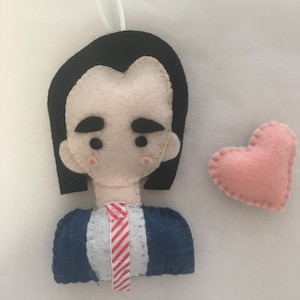 Puede incluir: Un adorno de fieltro de una persona con cabello negro, una chaqueta azul y una corbata a rayas rojas y blancas. Un corazón de fieltro rosa está a la derecha de la persona.