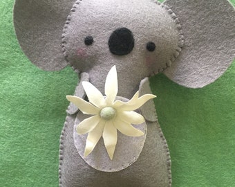 Può includere: Un koala di feltro grigio che tiene un fiore di feltro bianco. Il koala ha occhi neri e un naso nero.