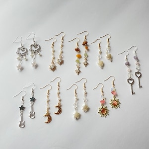 May include: Assortment of silver and gold earrings with various charms, including stars, moons, hearts, keys, and musical notes. The earrings are displayed on a white background.