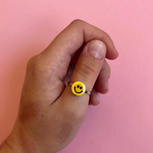 May include: A yellow smiley face ring with a black outline and a silver band. The ring is a fun and cheerful accessory. The ring is a small, round shape and is worn on a finger.