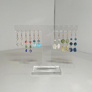 May include: A display of eight pairs of earrings with various colored beads and charms, including a butterfly, a flower, a crescent moon, and a star. The earrings are displayed on a clear acrylic stand.