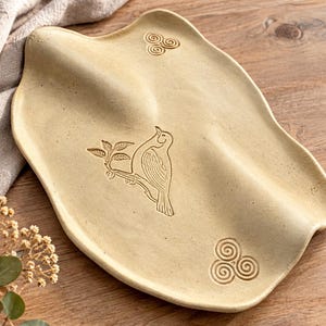 May include: A cream-coloured ceramic spoon rest with an irregular, organic shape. The surface features an embossed illustration of a bird perched on a leafy branch, flanked by two spiral triskelion symbols. The finish is matte and speckled.