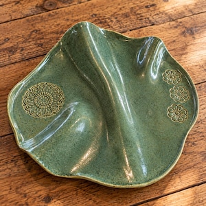 May include: A sage green ceramic serving dish with a wavy, organic edge and a central divider. The surface features a glossy, speckled glaze with a large circular floral stamp on the left side and three smaller flower stamps on the right side.