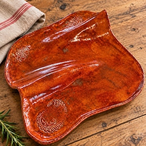 May include: A glossy, burnt-orange ceramic serving tray with an irregular, organic shape. The surface features a smooth, reflective glaze and two distinct embossed patterns, including a prominent paisley motif on the lower left section.