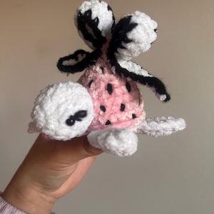 May include: A handmade crochet turtle with a pink shell dotted with black spots. The turtle has white head, feet, and a fluffy white and black wing-like structures. The turtle is held in a hand.