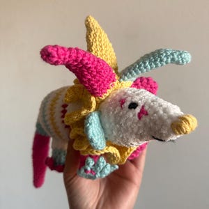 May include: A handmade crochet creature with a white body, yellow, pink, and blue accents. The whimsical design features a star-shaped marking on the face and a yellow snout. The creature has a colorful, layered headpiece and is held in a hand.