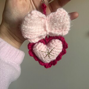 May include: A handmade pink crochet keychain featuring a bow and a heart. The bow is light pink with sparkly accents, and the heart is a lighter pink with a darker pink scalloped border and gold cross-stitch detail. A silver keyring is attached.