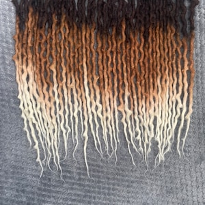 May include: A set of brown, blonde, and white ombre synthetic dreadlocks. The dreadlocks are long and wavy.