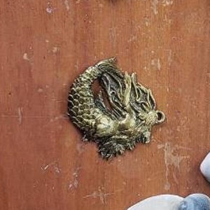 May include: A collection of seashells, a compass, a mermaid charm, and a piece of driftwood on a wooden surface. The seashells are in various shapes and sizes, and the compass is open and pointing north. The mermaid charm is made of metal and is in the shape of a mermaid with her tail curled around her.