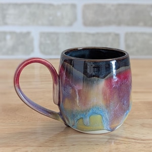 May include: A handcrafted ceramic mug with a vibrant glaze. The mug has a dark blue interior, a red handle, and a body with a mix of red, purple, blue, and yellow tones. The mug is placed on a wooden surface.