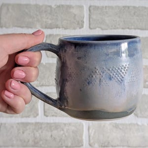 May include: A ceramic mug with a blue and grey glaze. The mug has a textured pattern and a large handle. The mug is held in a hand against a white brick wall. The mug is likely suitable for hot drinks.