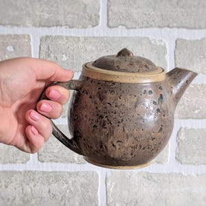 Peut inclure: Une théière en céramique marron avec un couvercle et une anse. La théière présente un motif tacheté et un bord marron clair. La théière est tenue par une main devant un mur de briques blanches.