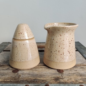 May include: A set of two ceramic cream and sugar containers with a speckled brown and white glaze. The containers have a light brown rim.
