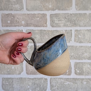 May include: A handmade ceramic mug with a unique design. The top half is glazed in a marbled blue, while the bottom half is a natural beige. The mug has a curved handle and is held against a brick wall.