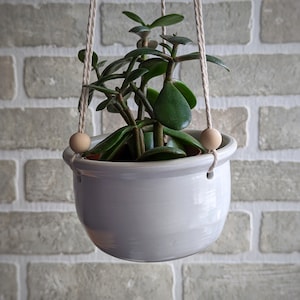 May include: A light grey ceramic hanging planter, suspended by natural rope. It contains a jade plant with green leaves. Wooden beads are used as decorative accents on the rope near the rim. The background is a light grey brick wall.
