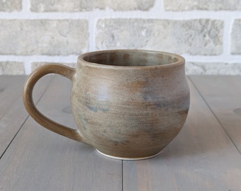 May include: A brown ceramic mug with a rounded body and a handle. The mug has a speckled glaze and a light brown color.