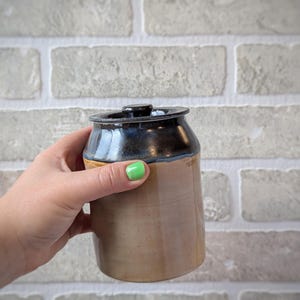 May include: A cylindrical ceramic jar with a smooth tan base and a dark blue glazed upper section. The jar features a matching ceramic lid with a small central knob handle, creating a two-toned appearance with a distinct horizontal color transition.