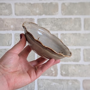 May include: A handmade, ceramic, leaf-shaped bowl with a wavy rim. The bowl is a muted grey colour with a brown rim. The bowl is held in a hand against a brick background. This decorative bowl is perfect for serving snacks or as a decorative piece.