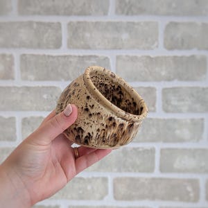 May include: A small, round ceramic bowl featuring a speckled beige and brown glaze. The exterior and interior surfaces display a textured, mottled pattern with dark brown spots concentrated towards the base and rim, creating a distinct organic appearance.