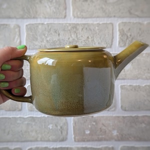 May include: A ceramic teapot with a glossy, mottled olive-green glaze featuring a subtle crackle texture. The teapot has a cylindrical body, a flat lid with a small central knob, and a short, angled spout extending from the side.