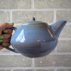 Peut inclure: Une théière en céramique avec une finition brillante dans des tons de bleu et de violet. La théière présente un corps arrondi, un bec verseur incurvé, une anse sur le côté et un couvercle assorti avec un petit bouton sur le dessus.