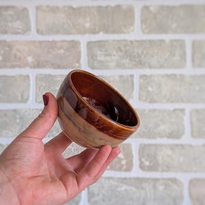 Peut inclure: Un petit bol en céramique fait à la main, de forme asymétrique unique. Le bol présente un dégradé de teintes marron et beige, avec une finition brillante. Le bol est tenu dans une main, mettant en valeur sa taille et son design.