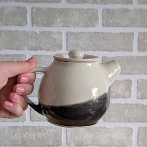 May include: A cream and dark grey ceramic teapot. The teapot has a small spout, a handle, and a lid with a small knob. The teapot is held by a person with pink nail polish. The teapot is likely to hold around 500 ml of liquid.