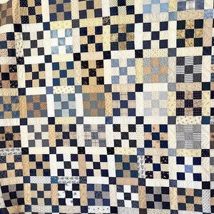 May include: A quilt made of various shades of blue, brown, and white fabric. The quilt is made up of small squares, each with a different pattern. The quilt is bordered with a dark blue fabric.