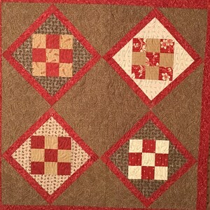 May include: A brown and red quilt with a diamond pattern. The quilt is made up of four smaller squares, each with a different pattern. The quilt is surrounded by a brown border.