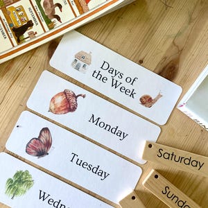 May include: A set of educational cards and wooden tags featuring the days of the week. The white cards display illustrations of a house, snail, acorn, butterfly, and tree alongside text. The wooden tags are engraved with the days of the week.