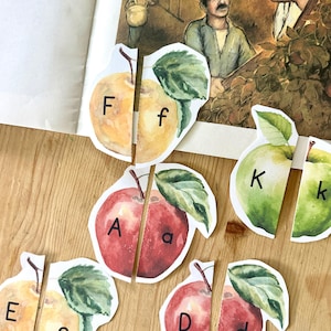 May include: A set of paper apple cutouts featuring watercolor illustrations of red, yellow, and green apples. Each apple is split into two pieces, with uppercase and lowercase letters F, f, A, a, K, k, E, e, D, and d printed on the fruit surfaces.