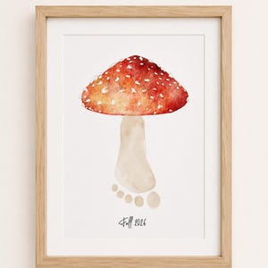 Könnte beinhalten: Ein gerahmter Aquarelldruck mit einem roten Pilzhut mit weißen Punkten auf einem beigen, fußabdruckförmigen Stiel. Das Kunstwerk enthält den Text "Fall 2026" in schwarzer Schreibschrift am unteren Rand, eingefasst in einen hellen Holzrahmen.
