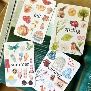 Può includere: Quattro carte rettangolari con illustrazioni ad acquerello che rappresentano le stagioni. Ogni carta mostra il testo fall, spring, summer o winter, circondato da icone tematiche come zucche, fiori, un faro e un camino.