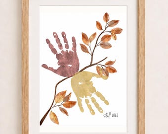 May include: A framed print featuring a brown handprint and a yellow handprint arranged around a central branch with orange and brown leaves. The text "Fall 2026" is written in black script in the bottom right corner of the white paper background.