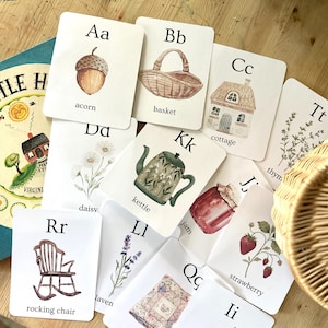 Op de afbeelding: Een verzameling witte educatieve alfabetkaarten met aquarelillustraties van voorwerpen zoals een eikel, mand, huisje, waterkoker, madeliefje, jam, aardbei, schommelstoel, lavendel en quilt, elk gelabeld met de bijbehorende letter.