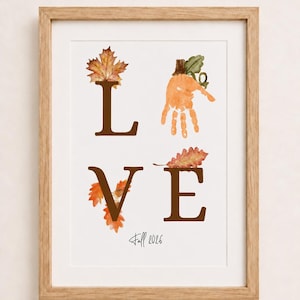 May include: A framed wall art print featuring the word LOVE in brown serif letters. The letter L is topped with a maple leaf, the O is an orange handprint with a pumpkin stem, the V is decorated with orange leaves, and the E has an oak leaf. Text reads Fall 2026.