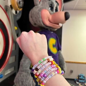 Peut inclure: Un poignet avec plusieurs bracelets en perles colorées. Les bracelets ont des perles de différentes couleurs et des perles en lettres qui épellent des noms, dont "Chuck E Cheese", "Jasper", "Isabella", "Bella", "Munch" et "Helen".