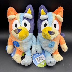 May include: Two plush toys of a cartoon dog with orange, blue, and yellow coloring. Each toy has a button on its chest and a tag attached. The toys are on a black surface.