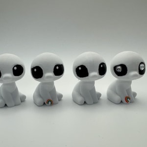 May include: Four small, white, alien-like figurines with large black eyes and small rainbow accents. One figurine has a sparkling gem in one eye. The figurines are sitting down.