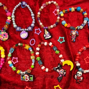 May include: A selection of colourful beaded bracelets and necklaces with various charms, including cartoon characters, stars, and other shapes. The jewellery is displayed on a red velvet surface.
