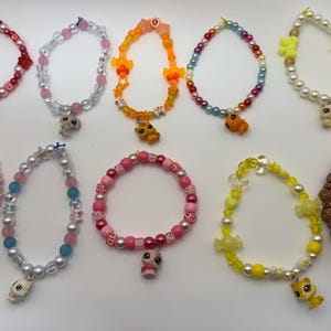 Puede incluir: Una colección de diez pulseras de cuentas en varios colores, incluyendo rosa, blanco, naranja y amarillo. Cada pulsera presenta un dije, como una estrella, un animal o una flor. Las pulseras están diseñadas para niños.