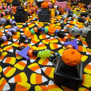 May include: A collection of beaded bracelets featuring Halloween-themed charms including orange jack-o'-lanterns, purple witch hats, and green ghosts. The bracelets are constructed with a variety of colorful plastic beads, including black, orange, purple, and iridescent spheres, arranged in circular patterns on black display stands.